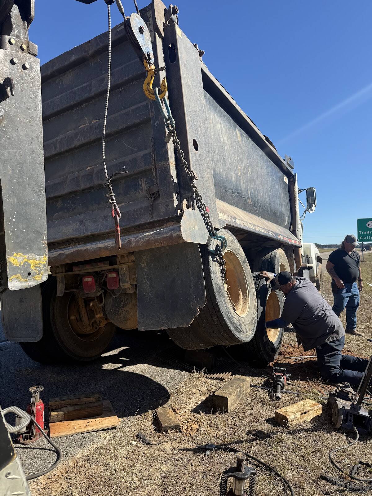 Two men working on the rear wheels of a dump truck held up by a wrecker's chains on a grassy highway shoulder