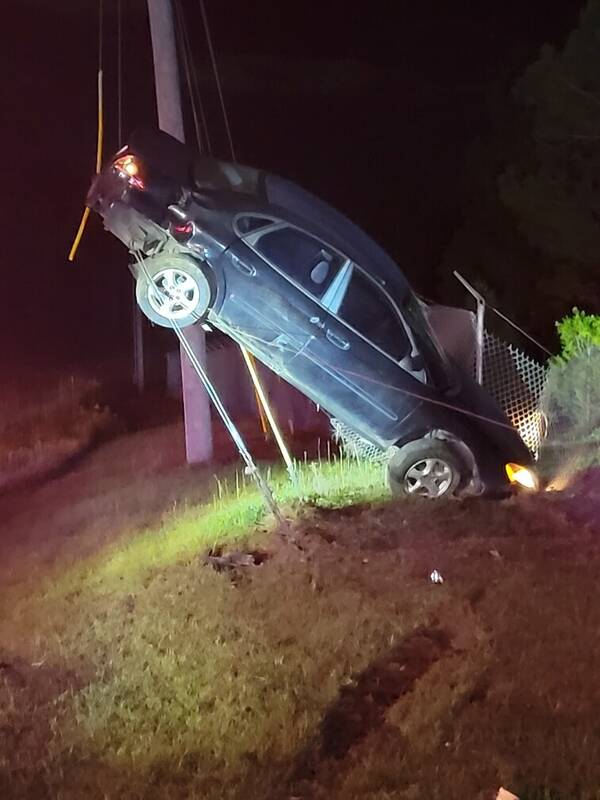 Dark sedan nose-down on a grassy embankment at night, rigged with straps under work lights