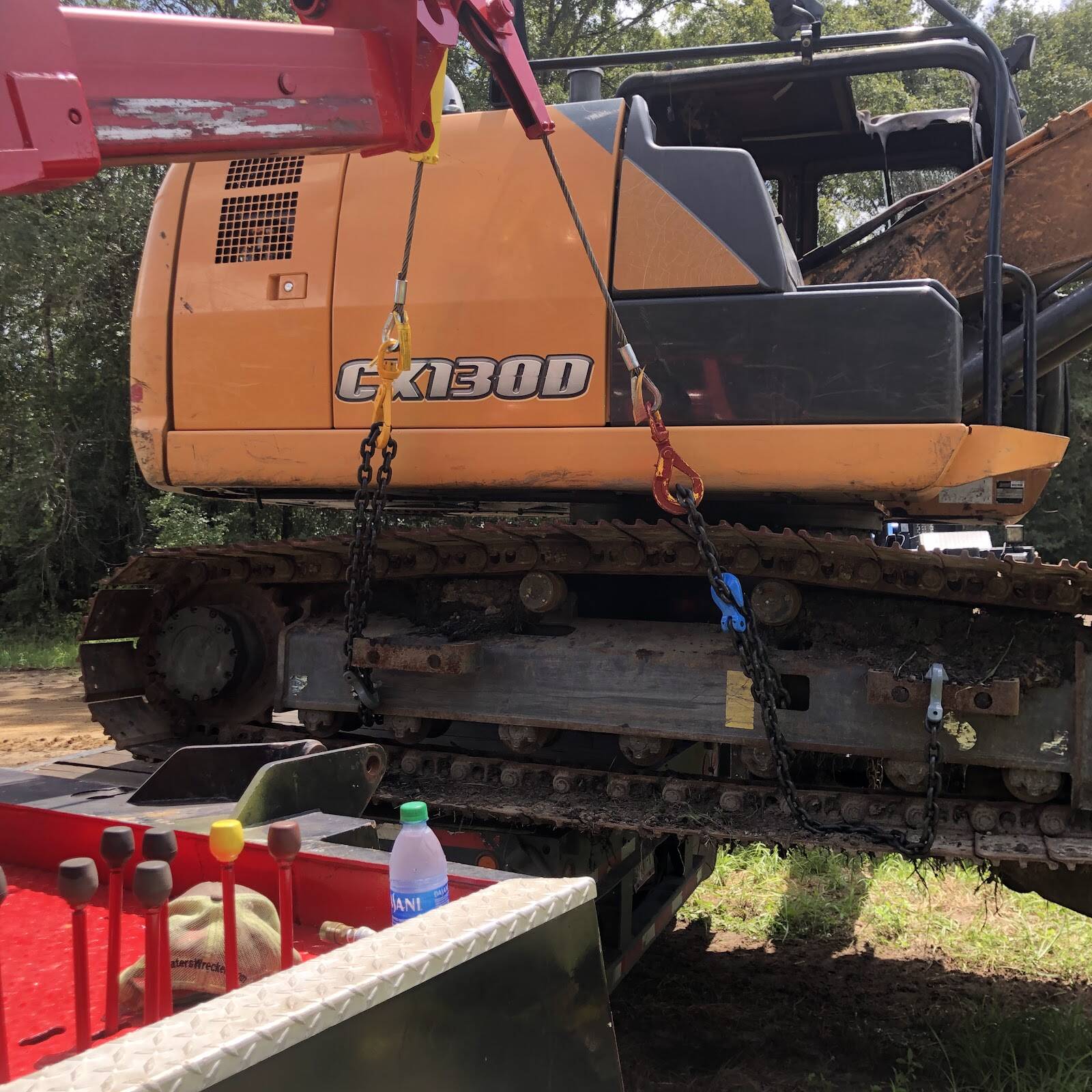 Red wrecker boom lifting an orange excavator by chains over a truck bed