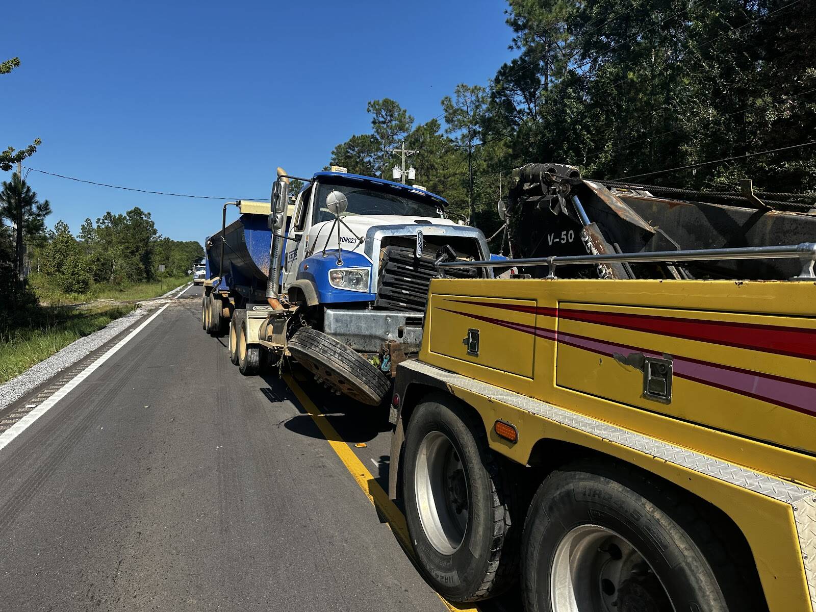 View from behind a yellow wrecker on a two-lane road through pines, a damaged blue semi with an end-dump trailer stopped ahead