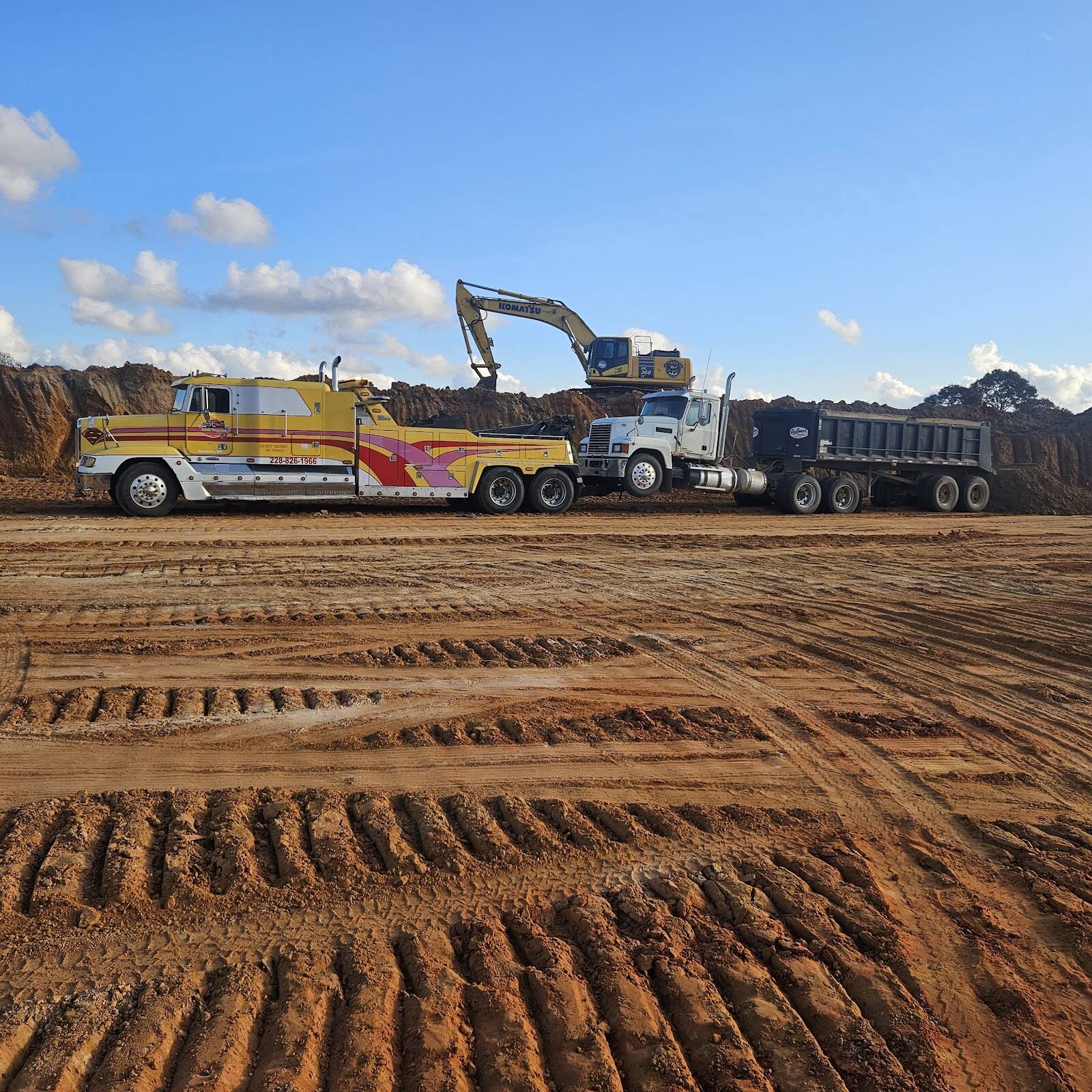 Yellow heavy wrecker with red and pink stripes hooked to a white dump truck and trailer on a red-dirt site, an excavator on the bank behind