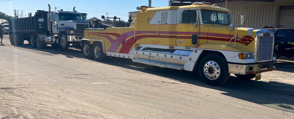 Yellow wrecker towing a white and blue Mack dump truck across a gravel lot beside a metal building